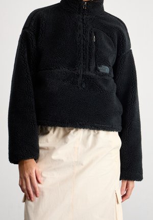 Pull polaire noir avec fermeture éclair quart de zip, poche poitrine et logo The North Face, porté avec une jupe beige clair.