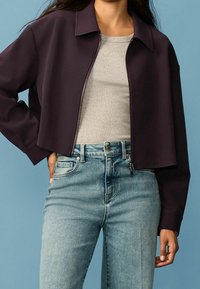 Kort lilla cropped jakke med krave og fuld lynlås, parret med en grå tætsiddende skjorte og lyseblå højtaljede jeans.