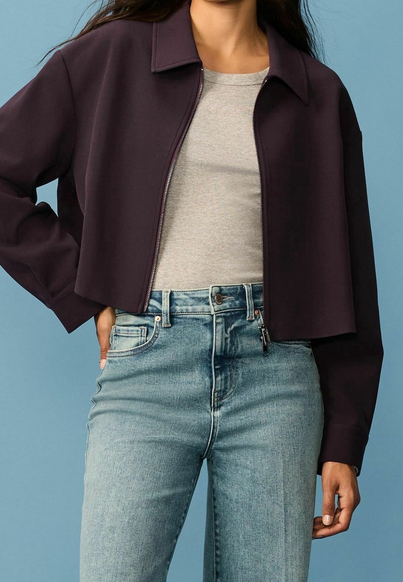 Kort lilla cropped jakke med krave og fuld lynlås, parret med en grå tætsiddende skjorte og lyseblå højtaljede jeans.