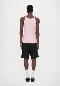 Lys rosa ermeløs tanktopp, svarte shorts, hvite sokker og svarte sko. Jevn tekstur og tettsittende design synlig fra baksiden.