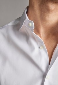 Close-up van de nek en borst van een persoon die een wit overhemd met knopen draagt, waarbij de bovenste knoop is losgemaakt.