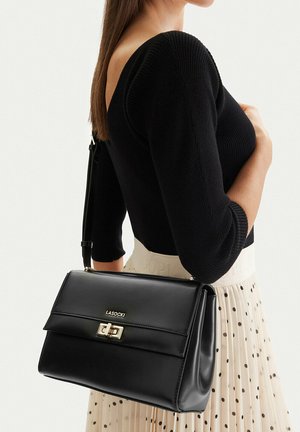 Donna che indossa un top nero a coste e una gonna beige a pois tiene una borsa nera Lasocki con chiusura dorata sulla spalla.