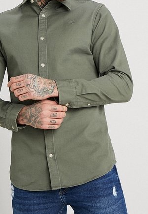 Man die een olijfgroen overhemd met knopen en blauwe versleten spijkerbroek draagt, met getatoeëerde handen die de mouw van het overhemd aanpassen.