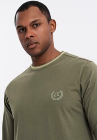 Olivegrünes Langarmshirt mit Rundhalsausschnitt, das auf der Brust die eingestickten Initialen "EX" in einem dekorativen Lorbeerdesign zeigt.
