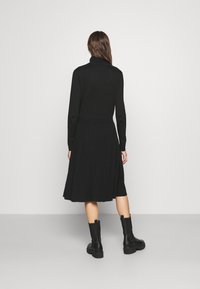Robes en maille côtelée noire avec de longues manches, col roulé et jupe plissée. Associée à des bottines noires épaisses. Vue de dos présentée.