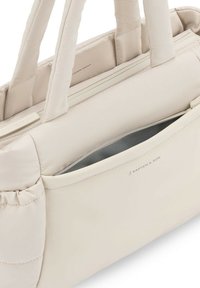 Sac beige doux rembourré avec poignées doubles, poche zippée avant partiellement ouverte, et logo Kapten & Son sur le devant de la poche.