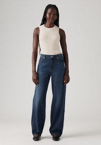 Hoge taille donkerblauwe denim jeans met een wijde broekspijp, gecombineerd met een crèmekleurige ribbelachtige mouwloze top. Gladde textuur, geen zichtbare patronen.
