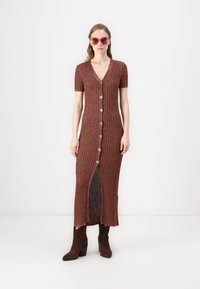 Braunes geripptes Maxikleid mit kurzen Ärmeln, Vorderknöpfung und einem Seitenschlitz. Kombiniert mit braunen Absatzstiefeln und übergroßen Sonnenbrillen.