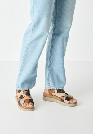Sandalias con tiras en una mezcla de tonos marrón, crema y negro, con una suela acolchada y diseño de punta abierta, combinadas con unos jeans de mezclilla claros.