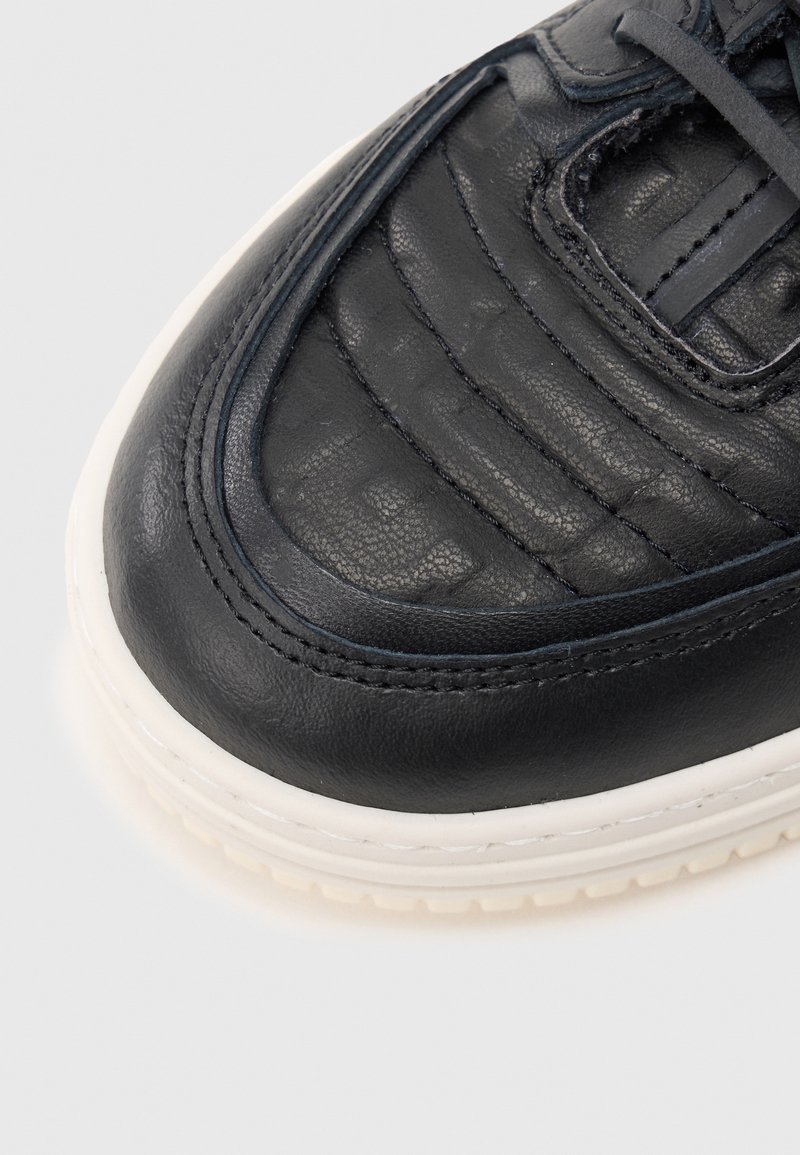 Black leather sneaker with a textured surface, tonal stitching, rounded toe, and a white rubber sole. Features subtle layered design accents.