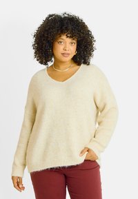 Pull en maille couleur crème avec un col en V, des manches longues et une texture douce. Associé à un pantalon bordeaux, offrant une coupe décontractée.