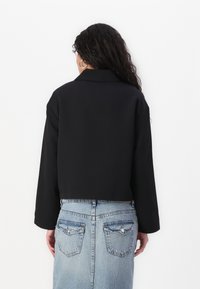 Veste noire courte avec un col cranté et des manches longues, en tissu lisse. Portée par-dessus une jupe en denim bleu clair.