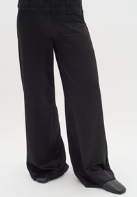 Pantalon noir à jambes larges fabriqué dans un tissu lisse et léger. Présente une coupe décontractée et un léger éclat, associé à des chaussures noires sans lacets.