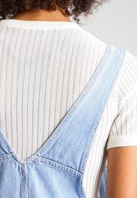 Blauwe spijkeroverall met brede banden over een witte geribbelde trui. De overall heeft een diepe V-hals aan de achterkant en zichtbare stikseldetails.