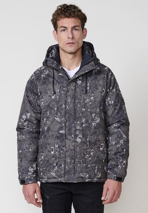 Chaqueta tipo puff en gris oscuro con un patrón abstracto blanco, con capucha, cierre de cremallera en la parte delantera y puños elásticos. Tejido aislante y texturizado.