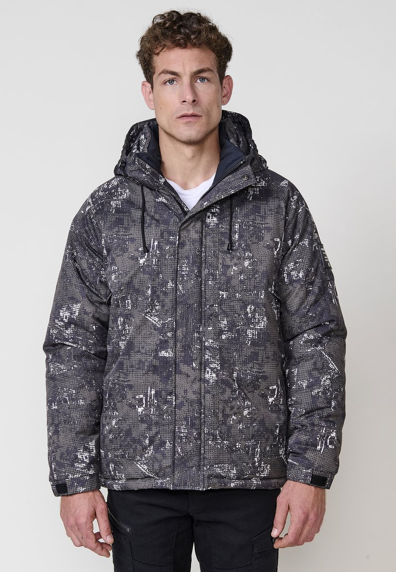 Chaqueta tipo puff en gris oscuro con un patrón abstracto blanco, con capucha, cierre de cremallera en la parte delantera y puños elásticos. Tejido aislante y texturizado.