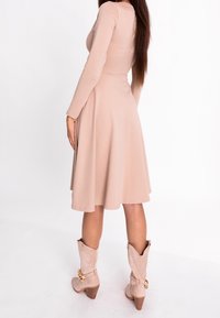 Robe à manches longues rose poudré avec un corsage ajusté, une jupe évasée et une texture douce ; associée à des bottines beiges ornées d'accents dorés.