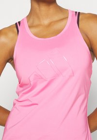 Różowy sportowy top na ramiączkach wykonany z gładkiej, elastycznej tkaniny. Na przodzie znajduje się błyszczące logo oraz warstwowe paski w kontrastujących kolorach.