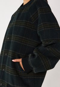 Giacca a quadri verde scuro con tessuto testurizzato, caratterizzata da una chiusura con bottoni, polsini a costine e tasche laterali decorate con dettagli neri.