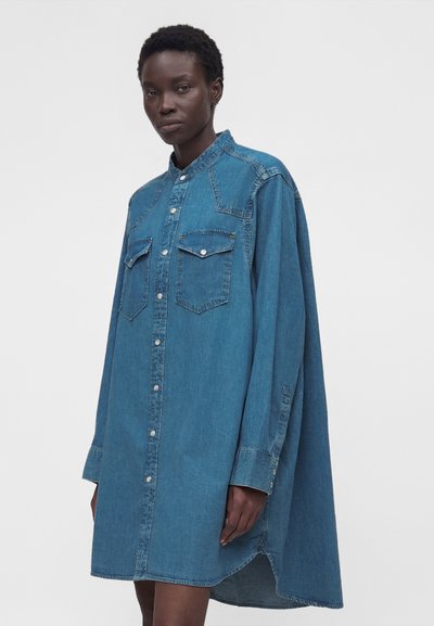 Γυναίκα που φοράει ένα φαρδύ, μακρυμάνικο μπλε τζιν φόρεμα πουκάμισο με κουμπιά και τσέπες στο στήθος, στέκεται μπροστά σε μια απλή, μονόχρωμη ταπετσαρία.