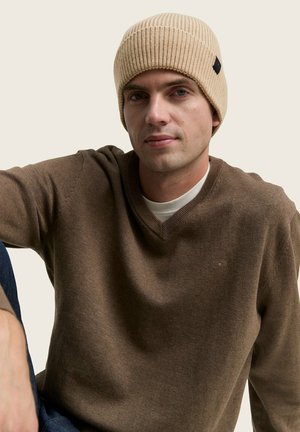 Beige knit beanie with ribbed texture. Model wears a brown V-neck sweater, sitting against a neutral background. Subtle branding visible.