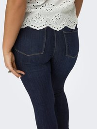 Nainen pukeutuneena tummansinisiin istuviin farkkuihin ja valkoiseen pitsipuseroon, kuvattuna takaa käsivarsi levitettynä lähelle reittä.