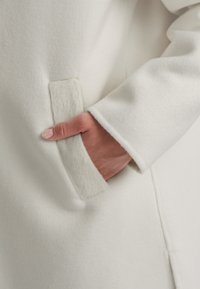 Weißer Wollmantel mit einer strukturierten, flauschigen Tasche. Glatte Ärmelkante. Nahaufnahme einer Hand, die in der Tasche ruht, betont die Stoffdetails.
