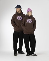 Sudaderas marrones con diseño gráfico rosa llevadas por dos personas. Pantalones anchos negros, zapatos casuales negros y un gorro rosa en uno de los individuos.