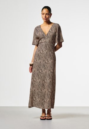 Vrouw draagt een lange V-hals jurk met zebraprinten, korte mouwen, sandalen en een armband, staand tegen een effen witte achtergrond.