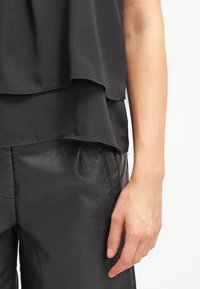 Bras droit et main d'une personne portant un haut noir superposé et un pantalon en cuir noir sur fond blanc.