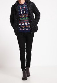 Zwarte gewatteerde jas over een kleurrijke kersttrui met Santa, pinguïns en bomen, gecombineerd met zwarte broek en grijze enkellaarsjes.