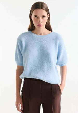 Vrouw met steil bruin haar die een lichtblauwe, zachte trui met korte mouwen en donkerbruine broek draagt, staand met één hand in de zak.