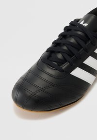Chaussure de football en cuir noir avec des rayures blanches, surface texturée, bout rond et lacets noirs. Semelle en caoutchouc visible au bas.