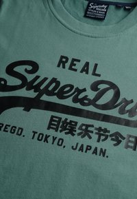 Zielona koszulka z czarnym napisem "Real SuperDry", japońskimi znakami oraz napisem "Regd. Tokyo, Japan" na przodzie.