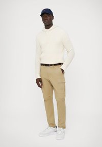 White textured hoodie, beige cargo pants with side pockets, and white sneakers. Blue cap with logo accent. Simple, casual outfit.