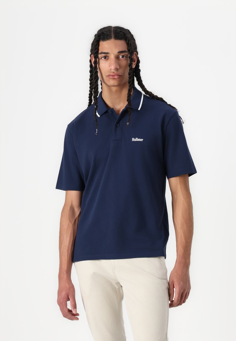 Mornarska polo majica z belimi ovratniki, kratkimi rokavi in vezenim logotipom na prsih. Model nosi svetle hlače.