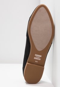 Semelle d'une chaussure TOMS noire avec semelle en caoutchouc marron, surface antidérapante texturée, marquage taille W6, inclinée contre une surface blanche.