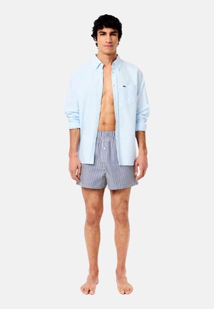 Hellblaues Button-Down-Hemd mit kurzen Ärmeln, offen über gestreiften Badehosen getragen. Die Badehosen haben horizontale Streifen in Marineblau und Weiß.