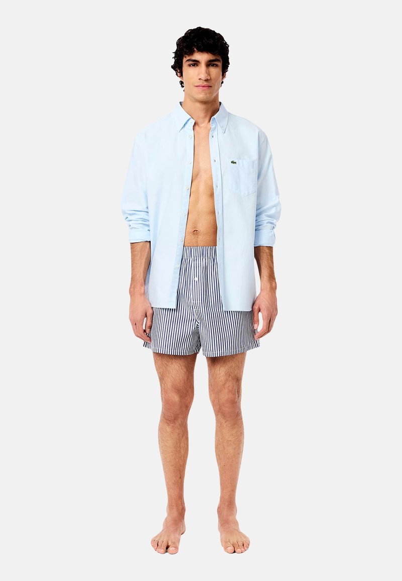 Light blue button-up shirt, short-sleeved, worn open over striped swim trunks. Trunks feature horizontal stripes in navy and white.