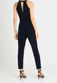 Mörkblå jumpsuit med figursydd silhuett, ärmlös design, öppen ryggdetalj och ankellånga, avsmalnande ben. Accepterad med svarta klacksandaler.