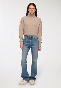 Beżowy sweter z wysokim kołnierzem, krótki fason; jasnoniebieskie dżinsy o wysokiej talii z rozszerzanymi nogawkami; kremowe sneakersy z teksturowanym wykończeniem.