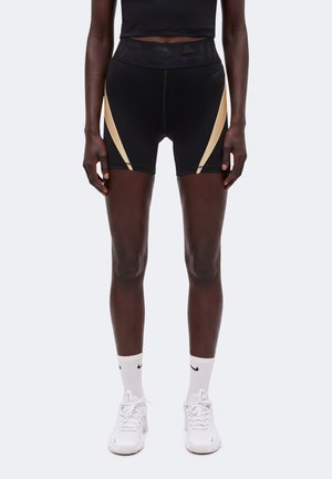 Person iført sorte træningsshorts med beige sidestriber, hvide crew-sokker og hvide sneakers, stående mod en ensartet lys baggrund.