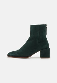Botines de ante verde oscuro con punta afilada y tacón cuadrado; cuenta con un cierre de cremallera lateral; textura suave y diseño minimalista.