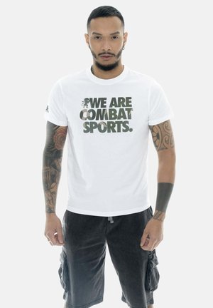Uomo con tatuaggi su entrambe le braccia, indossa una maglietta bianca con la scritta "WE ARE COMBAT SPORTS." e pantaloncini cargo neri, in piedi con il volto rivolto in avanti.