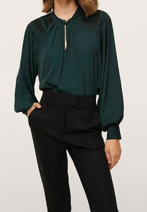 Kobieta ubrana w ciemnozieloną bluzkę z długim rękawem i plisowanymi ramionami oraz czarne, dopasowane spodnie z wysokim stanem, z jedną ręką w kieszeni.