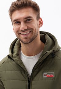 Giacca puffer khaki con cappuccio, dotata di chiusura zip, polsini a coste e una patch con un design rosso e grigio sul petto.