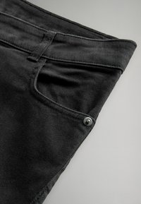 Tessuto di denim nero piegato con una tasca frontale, cuciture visibili e un dettaglio con bottone argentato sulla tasca per una chiusura sicura.