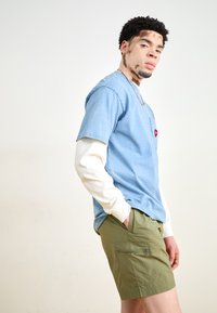 Camisa de mezclilla azul de manga corta superpuesta sobre una camisa blanca de manga larga, combinada con pantalones cortos verdes con un bolsillo. El modelo está de lado.