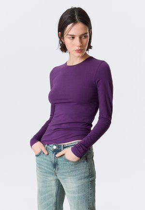 Moteris su trumpais tamsiais plaukais, dėvinti aptemptą violetinį ilgomis rankovėmis marškinėlį ir šviesiai mėlynas džinsus, stovinti su rankomis kišenėse, žvelgianti į šoną.