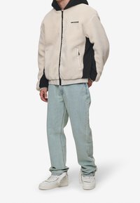 Giacca in pile di colore crema e nero, con zip frontale, cappuccio, bordo elastico in vita e tasche laterali. Indossata con jeans azzurri e sneackers bianche.